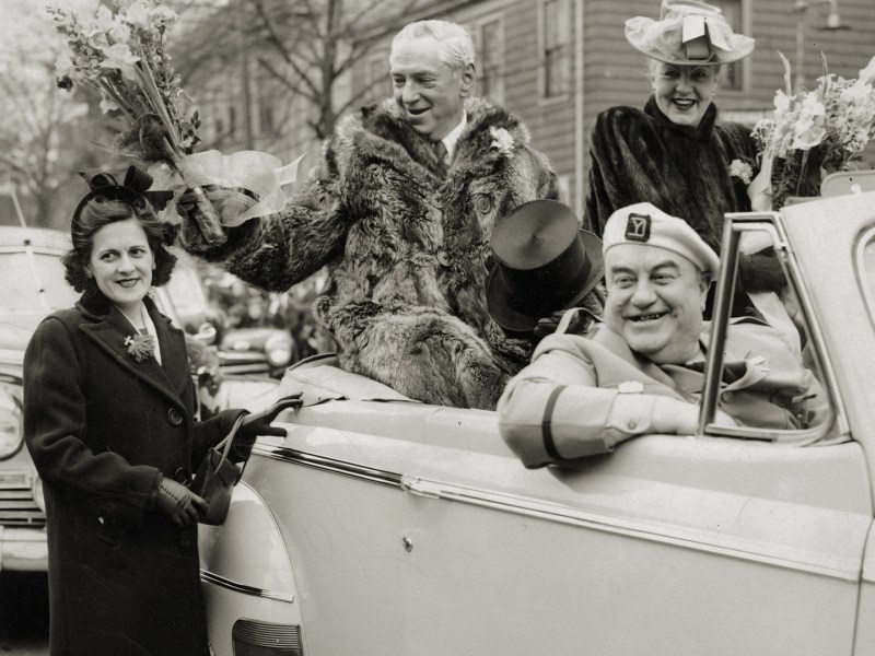 Photo of Mayor James Michael Curley, dressed in his raccoon coat, hands out flowers during South Boston's traditional Evacuation Day parade on March 17, 1947. Mayor Curley's wife, Gertrude, in a smart green hat with a pink ribbon is sitting to his left and Edward J. "Knocko" McCormack in his Yankee Division uniform is in front. The parade originally commemorated the day the British left Boston on March 17, 1776 and now it also honors St. Patrick, the patron saint of Ireland.