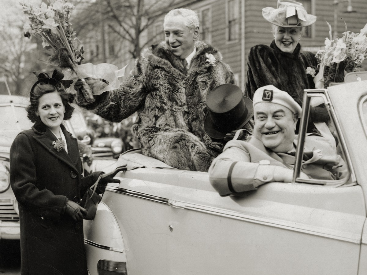 Photo of Mayor James Michael Curley, dressed in his raccoon coat, hands out flowers during South Boston's traditional Evacuation Day parade on March 17, 1947. Mayor Curley's wife, Gertrude, in a smart green hat with a pink ribbon is sitting to his left and Edward J. "Knocko" McCormack in his Yankee Division uniform is in front. The parade originally commemorated the day the British left Boston on March 17, 1776 and now it also honors St. Patrick, the patron saint of Ireland.