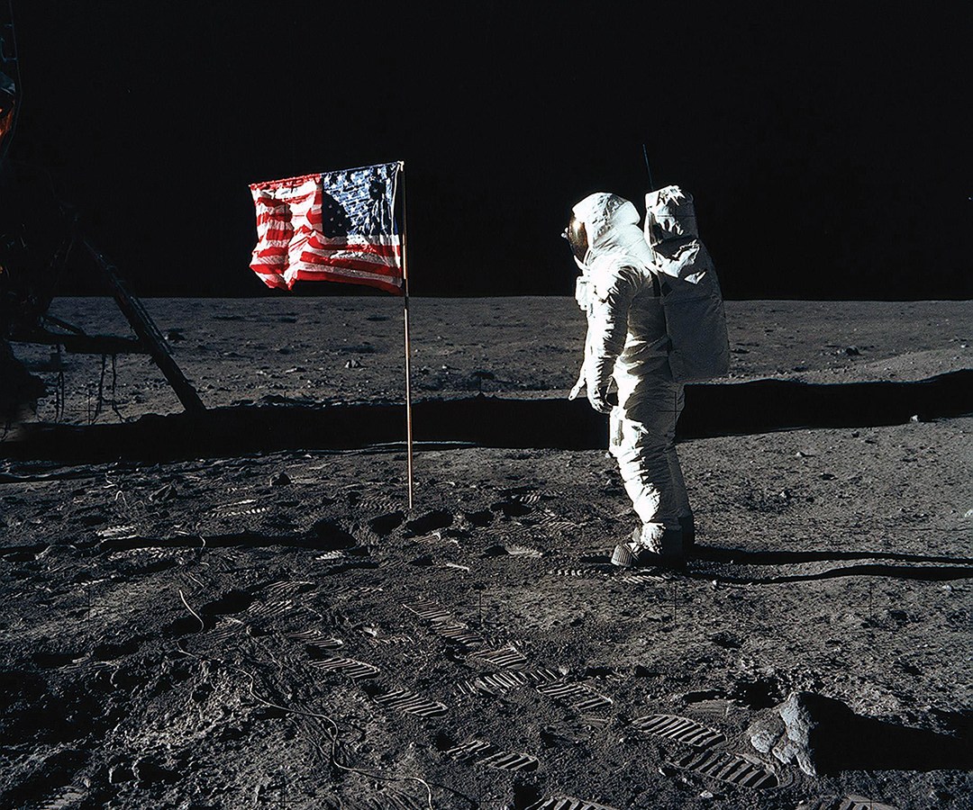 Photo of astronaut Edwin E. Aldrin Jr., lunar module pilot of the first lunar landing mission, poses for a photograph beside the deployed United States flag during an Apollo 11 extravehicular activity (EVA) on the lunar surface. The Lunar Module (LM) is on the left, and the footprints of the astronauts are clearly visible.
